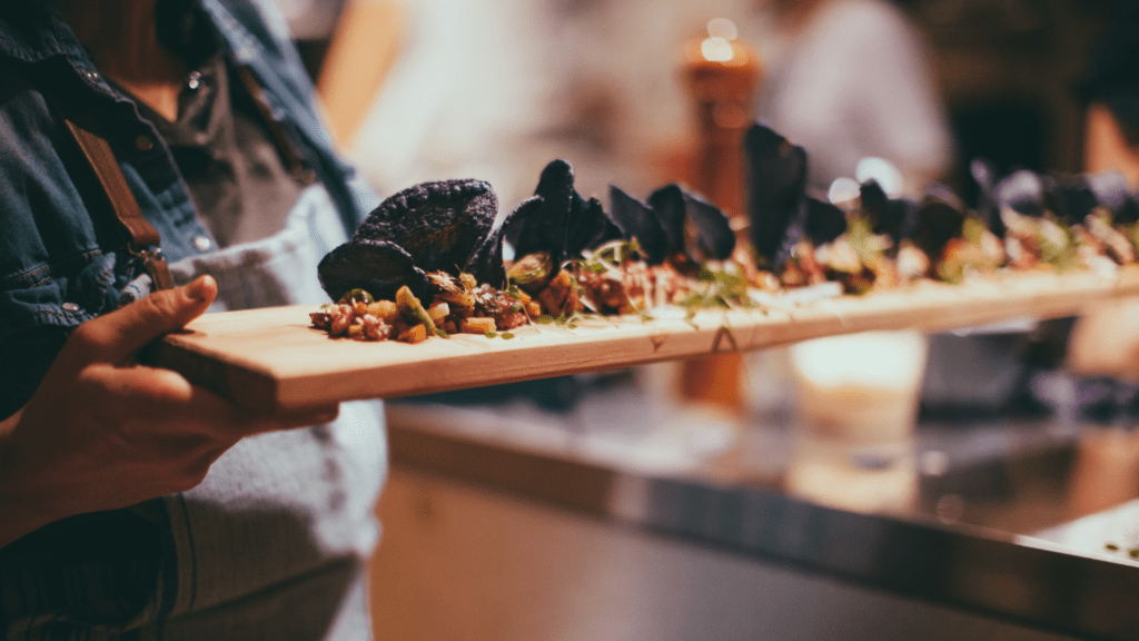 A professional caterer stands near an elegantly decorated event table, holding a tablet displaying catering and event planning software. The screen shows a client’s event itinerary, guest list, and customized menu. The setting suggests a wedding or upscale corporate event. This image emphasizes how catering software helps streamline planning, even while onsite.